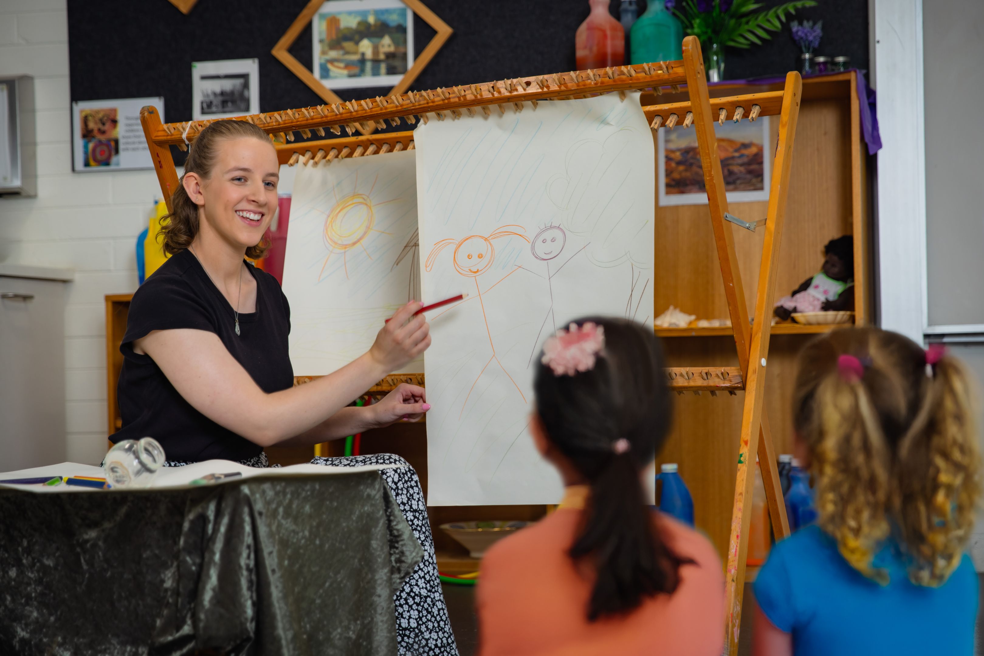An early childhood educator sits beside an easel, drawing simple figures with crayons while a group of children sits on the floor watching.