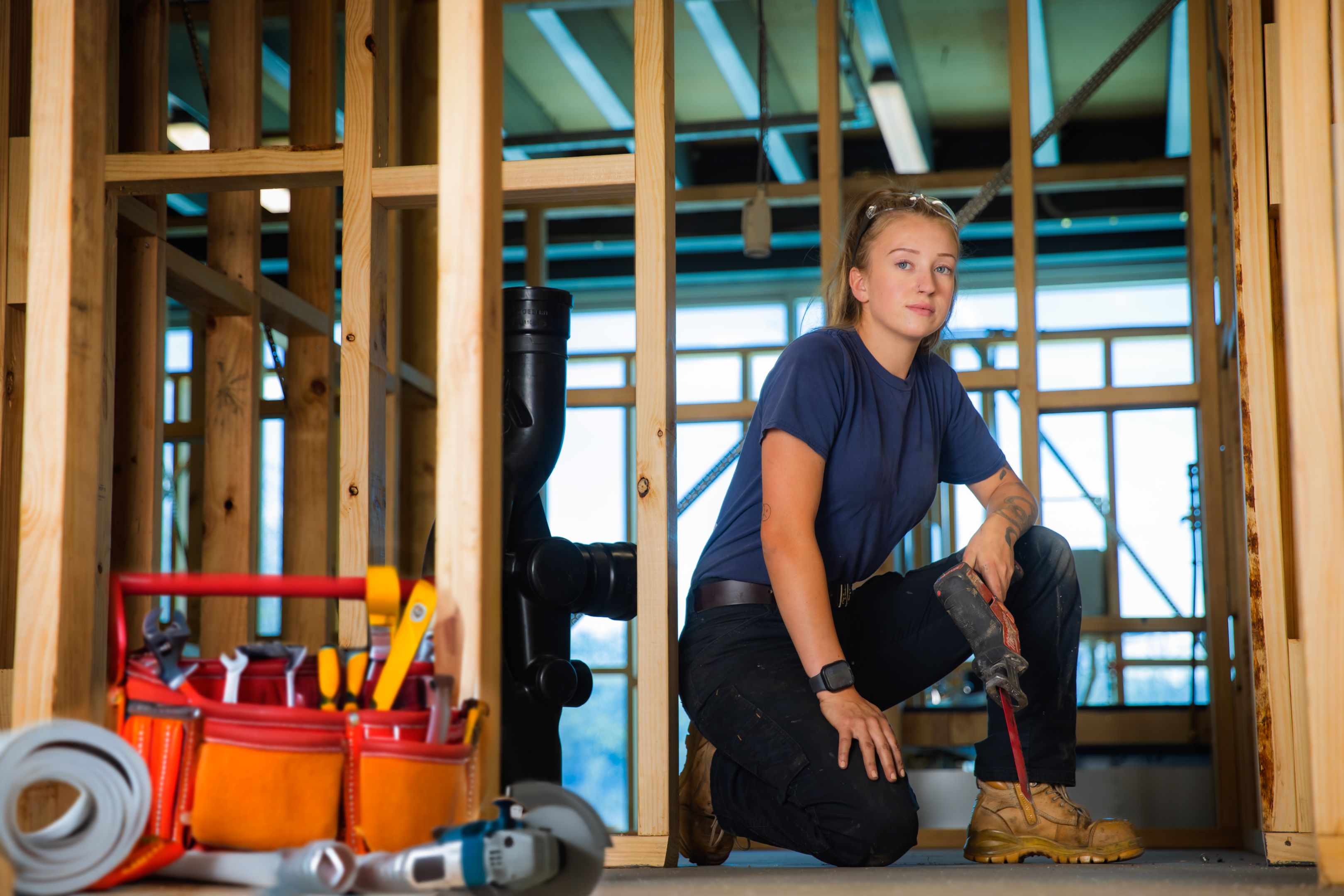 A young female construction worker kneels inside a partially framed building, holding a power drill with a tool bag and various equipment nearby.