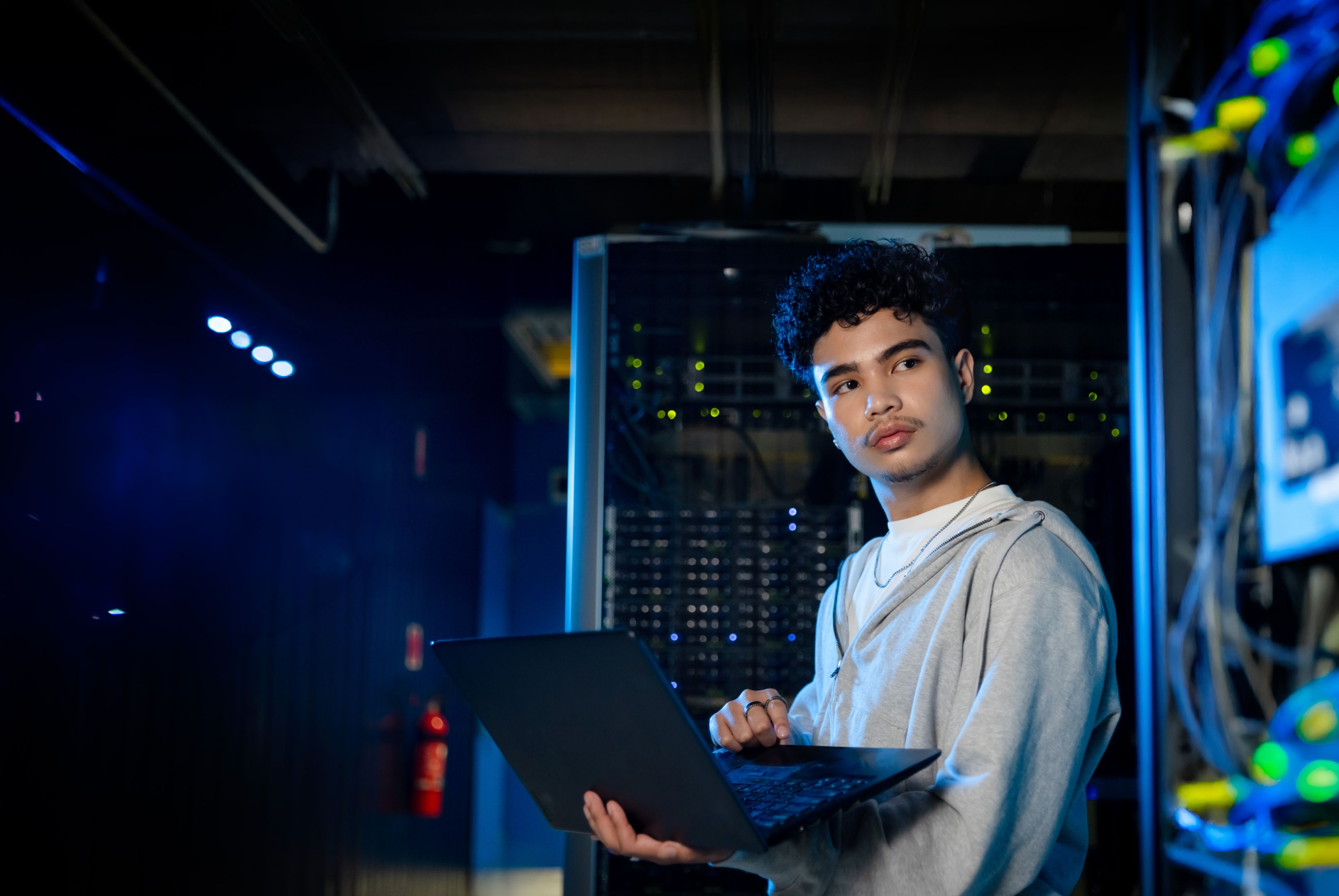 A young male student uses a laptop in a dimly lit server room, surrounded by racks of networking equipment and glowing indicator lights.