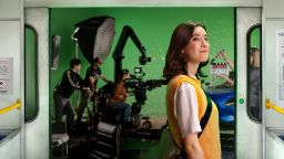 Young woman standing on a train as doors open to reveal a film set behind her.