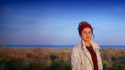 A young woman in a tan jacket, stares off in to the distance on a coastline.
