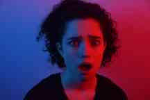 Woman with dark hair looking surprised at the camera with blue and red lights shining on her.