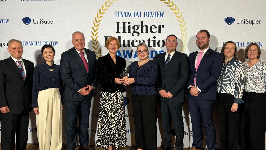 Nine people stand in front of a media wall reading 'Financial Review Higher Educations Awards'. At the centre, Swinburne Vice-Chancellor and President Professor Pascale Quester and Embolyability Director Professor Ruth Bridgstock hold a trophy.