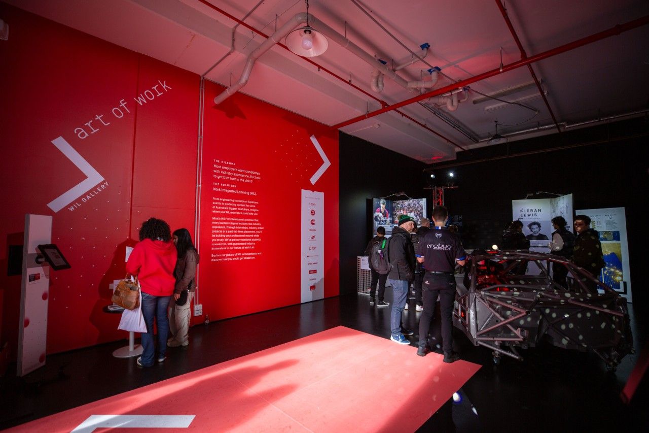 The WIL Gallery with a statement red wall, red carpet entrance and guests engaging with the featured WIL students.