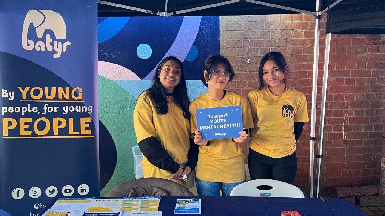 Three batyr student ambassadors at an on-campus stall, with one holding a sign that says “I support youth mental health”.