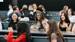 Smiling students sitting in a lecture theatre. One in the middle is waving.
