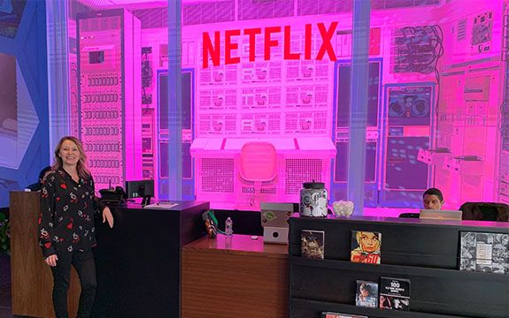 Swinburne alum Sue Bolton profile Sue wears a black shirt and pants and stands in front of the Netflix reception areas. A huge desk and chair installation can be seen behind a glass wall and the space is illuminated by pink neon light.