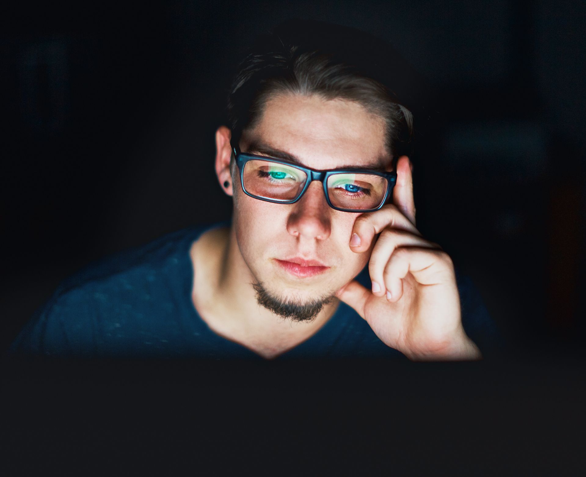 A young man in glasses, lit only by his computer monitor, stares at the screen. Young man sits in the dark staring at computer monitor