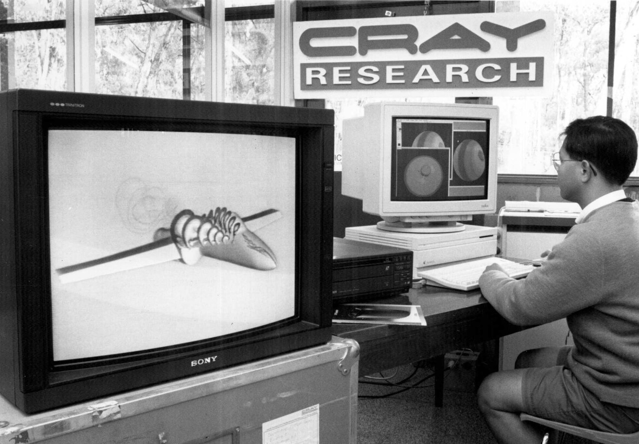 A black and white photograph of a man sitting at a '90s style computer. He is looking at graphics on screen