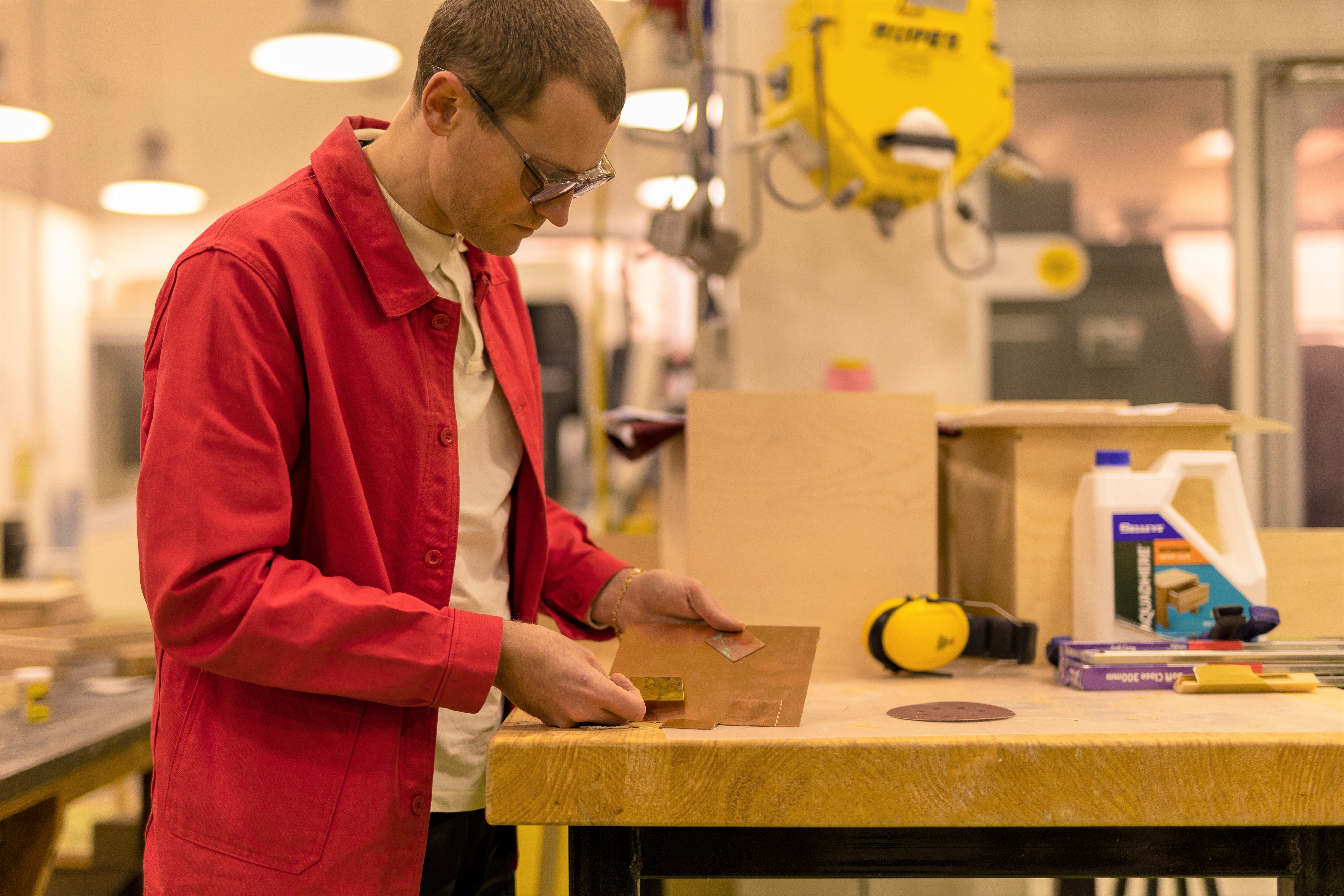 A man in a red coat and glasses studies samples of copper on a workbench surrounded by equipment 
