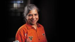 A female with greying hair is wearing an orange shirt and stands against a dark background.