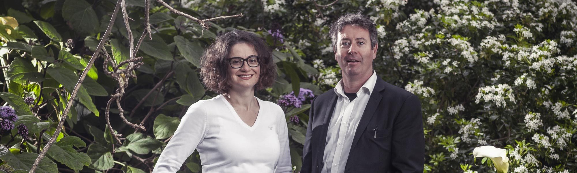 Professor Niki Frantzeskaki & Dr Stephen Glackin Professor Niki Frantzeskaki wearing a white top on the left and Dr. Stephen Glackin wearing a dark blazer on the right, both pictured in front of a luscious green garden.