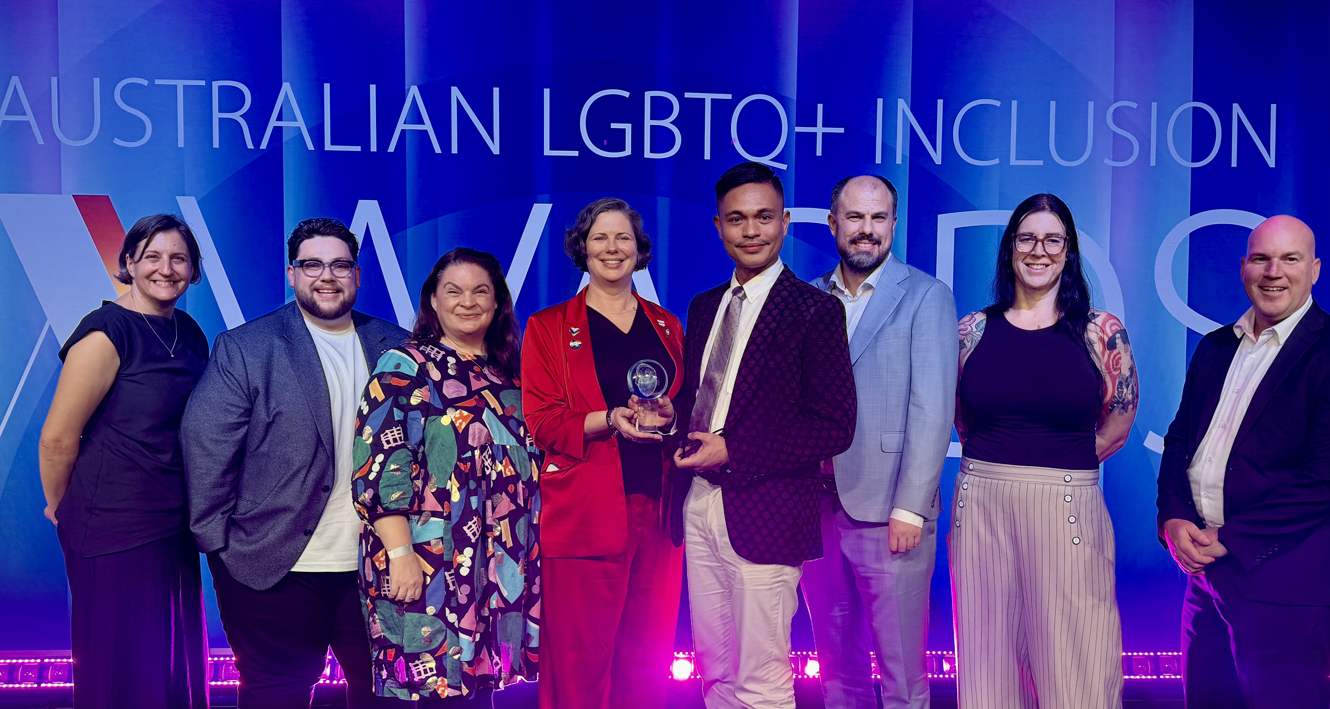 Group of people standing in front of a background that says "Australian LGBTQ+ inclusion", holding a trophy
