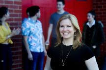 Young blonde woman in a black t-shirt smiling at the camera, group of people in the background behind her blurred out.