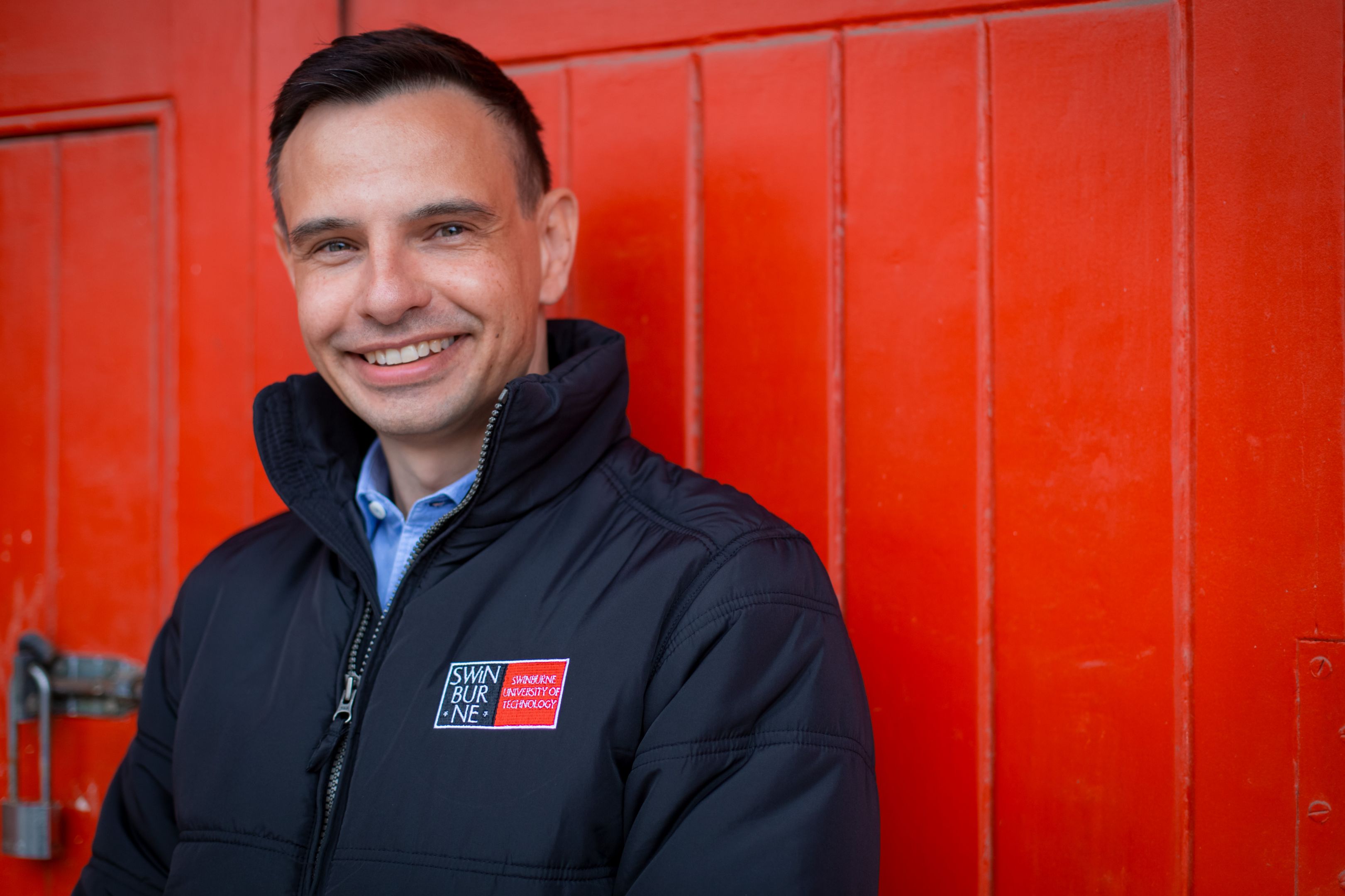 Werner van der Merwe Werner smiles to camera wearing a black jacket featuring the SUT logo with a bright red wall in the background