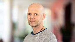 Bald man in a grey sweater standing slightly side on, looking at the camera.