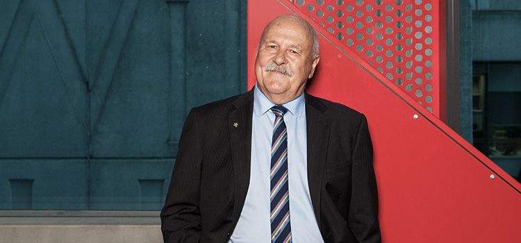 Associate Professor Bruce McDonald An older man dressed in a suit stands in front of a navy and red building
