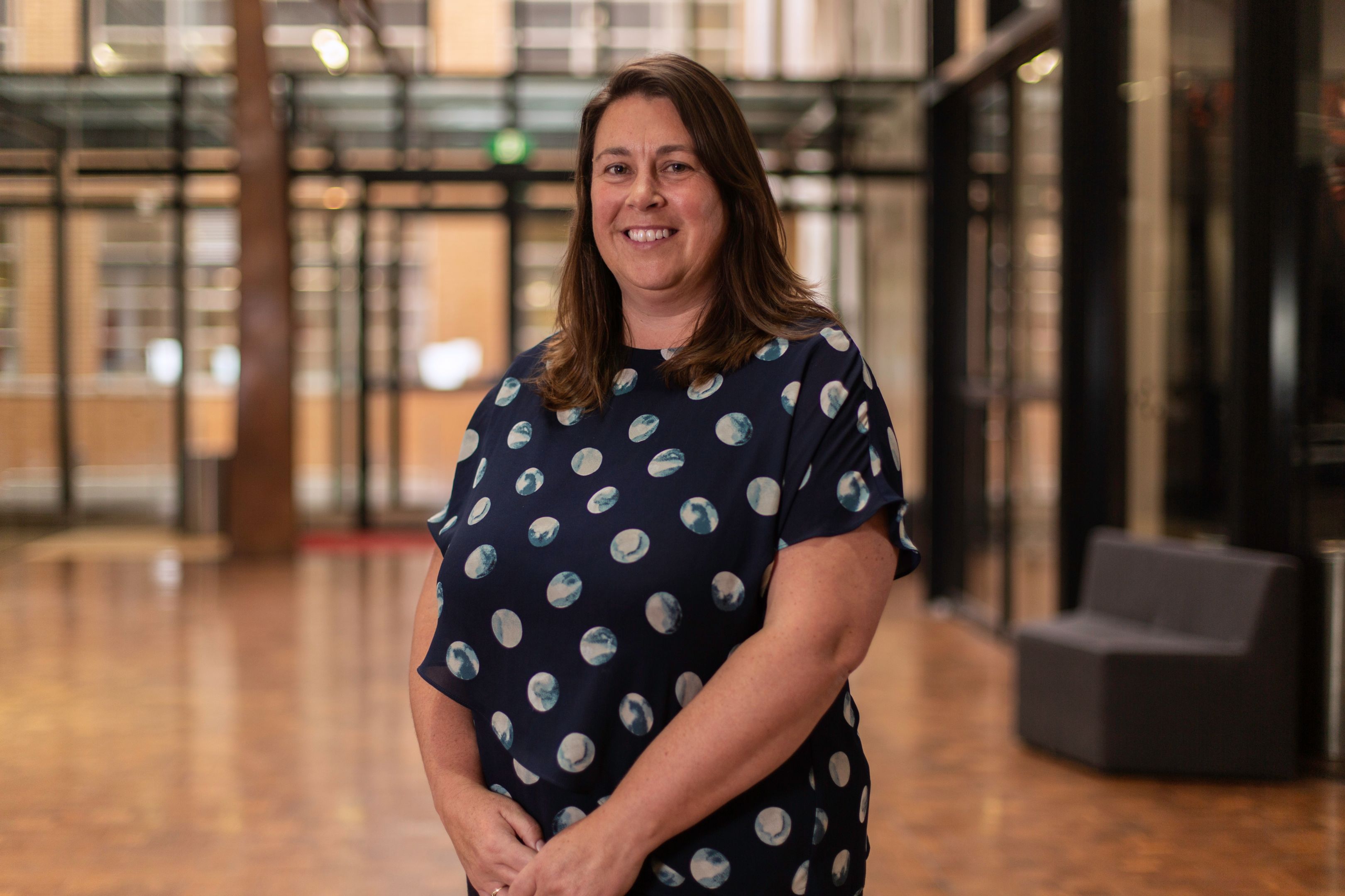 Megan Hough, Collaborations and Partnerships Megan Hough poses with hands clasped out in front wearing a patterned navy blue dress in the AMDC building