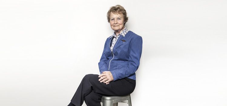 Dr Judith Kinnear An older woman dressed in a blue jacket and black pants sits in a relaxed pose on a stool