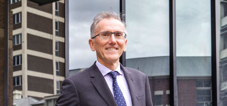 Professor Michael Gilding A man dressed in a suit stands in front of buildings in the city and smiles at the camera
