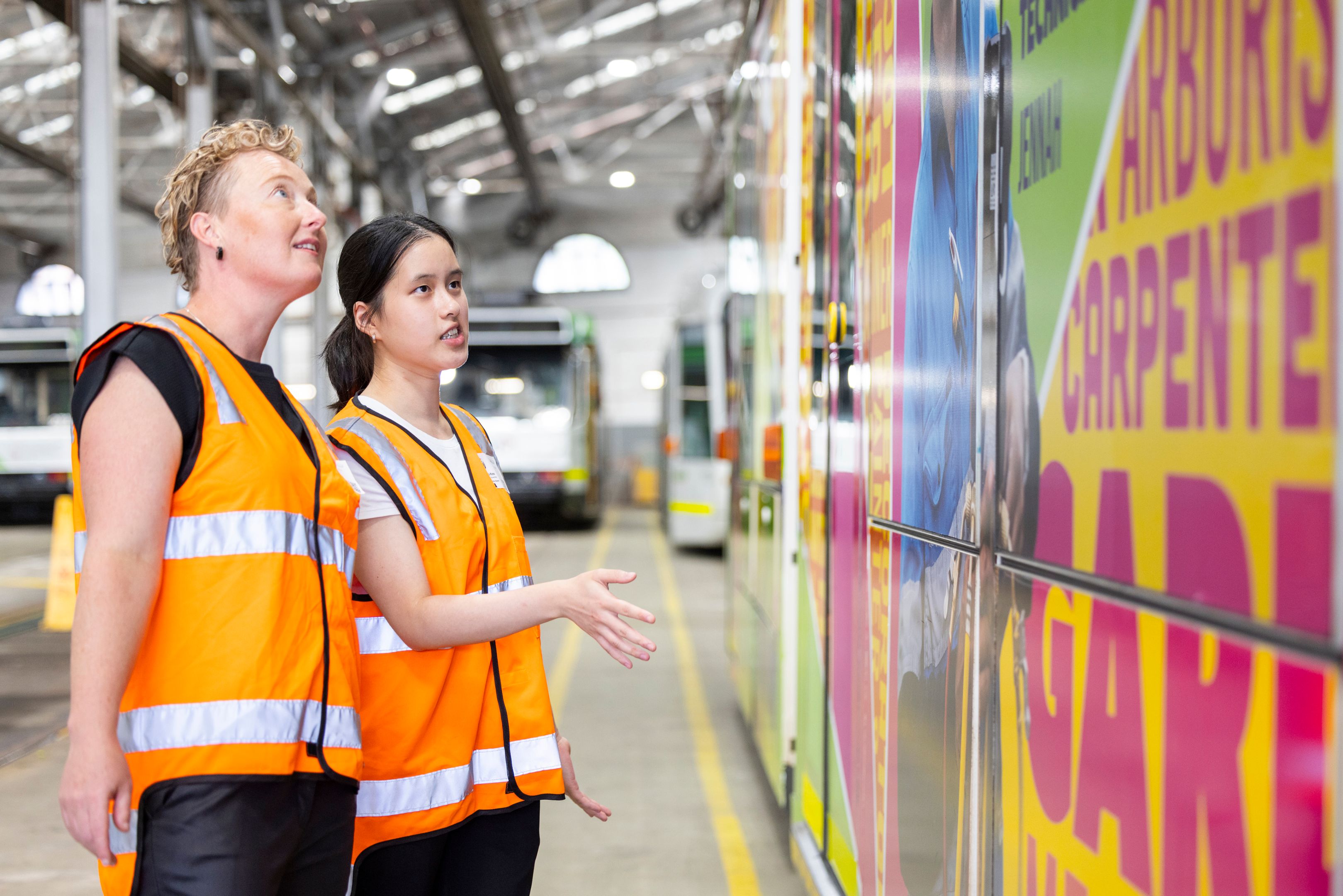 Two women in orange hi-vis vests in a tram warehouse looking at the colourful artwork on a tram