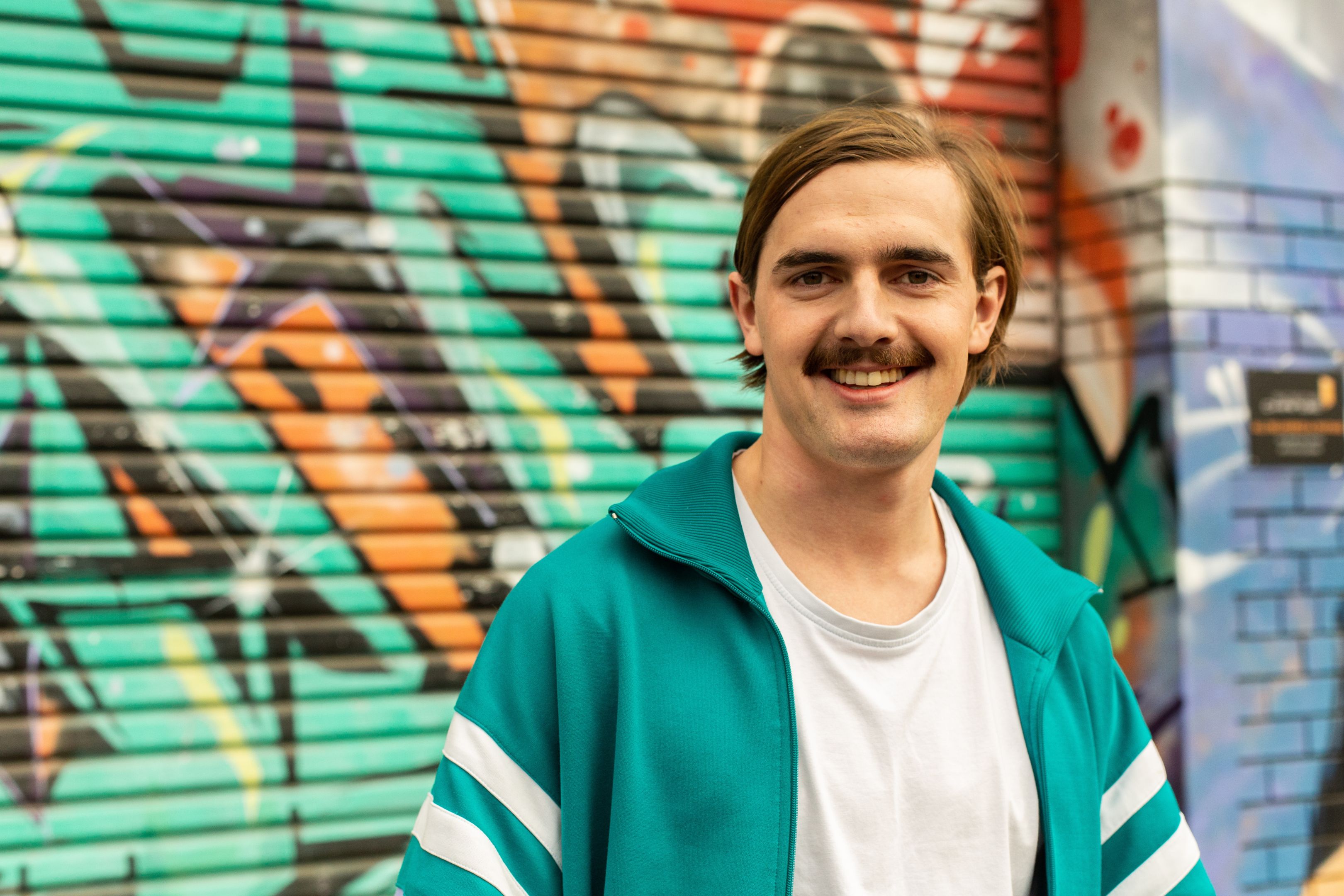 Man with stache standing in front of a graffiti-ed wall, looking pleased with his life choices. Man with stache standing in front of a graffiti-ed wall, looking pleased with his life choices.