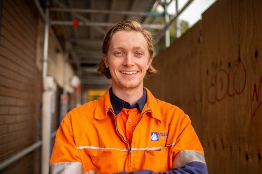 Jacob Stanford A smiling young man wearing an orange high-vis work shirt stands in front of a construction site