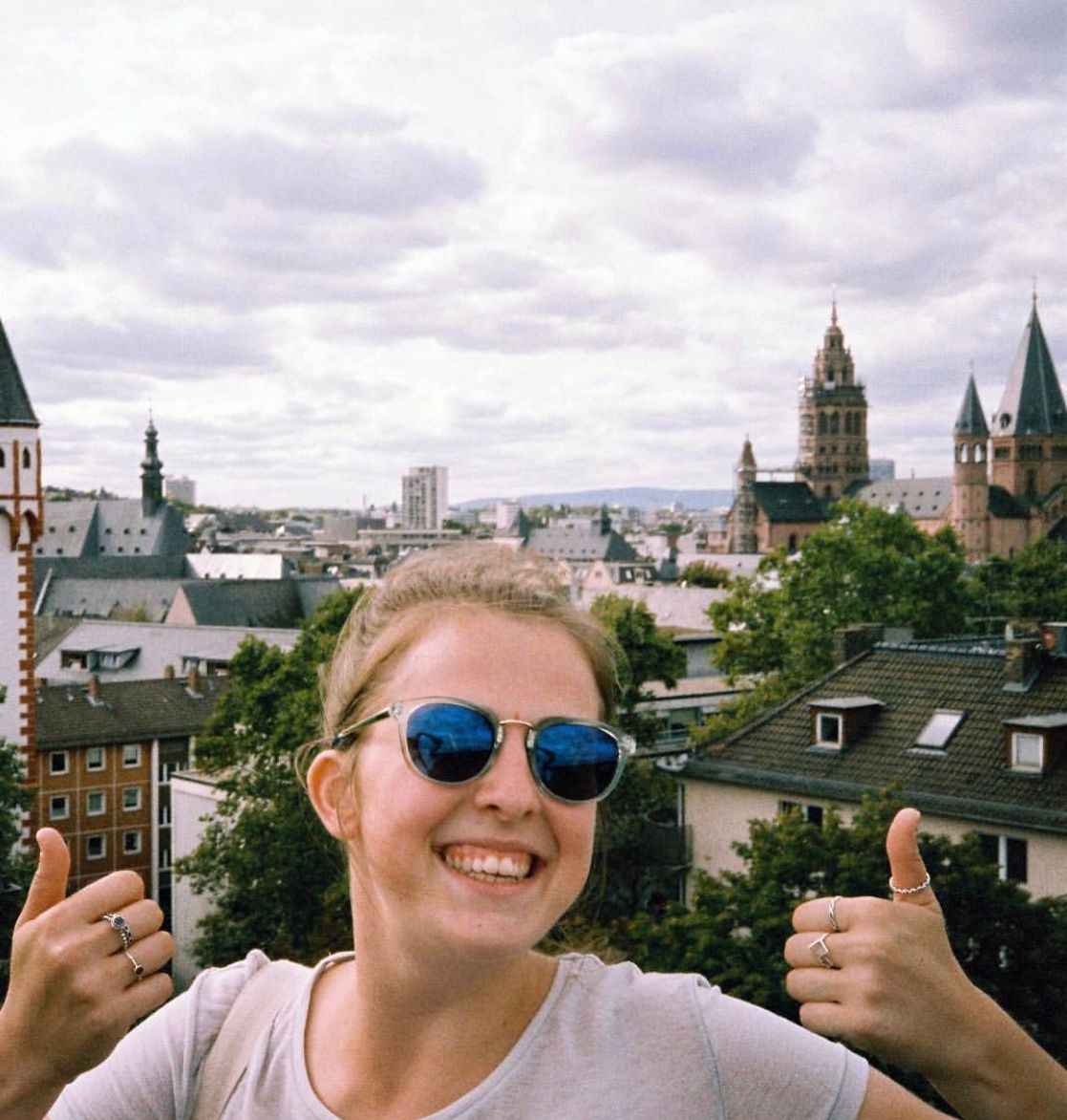 Study abroad and exchange student Maddie Female smiling at the camera with both thumbs up and the Germany city scape in the background