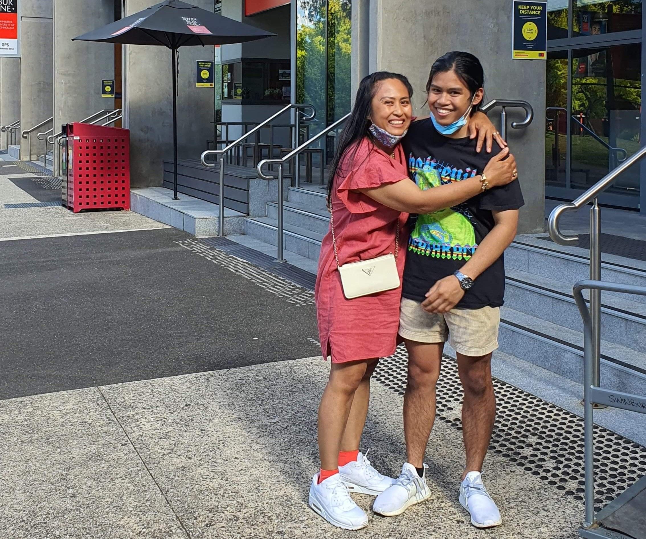 Swinburne Bachelor of Aviation / Bachelor of Business student Stephen (right) Swinburne student Stephen wears summer clothing and stands outside the concrete steps and glass doors to Swinburne student residences with his mum. His mum is hugging him and they both are smiling happily at the camera.