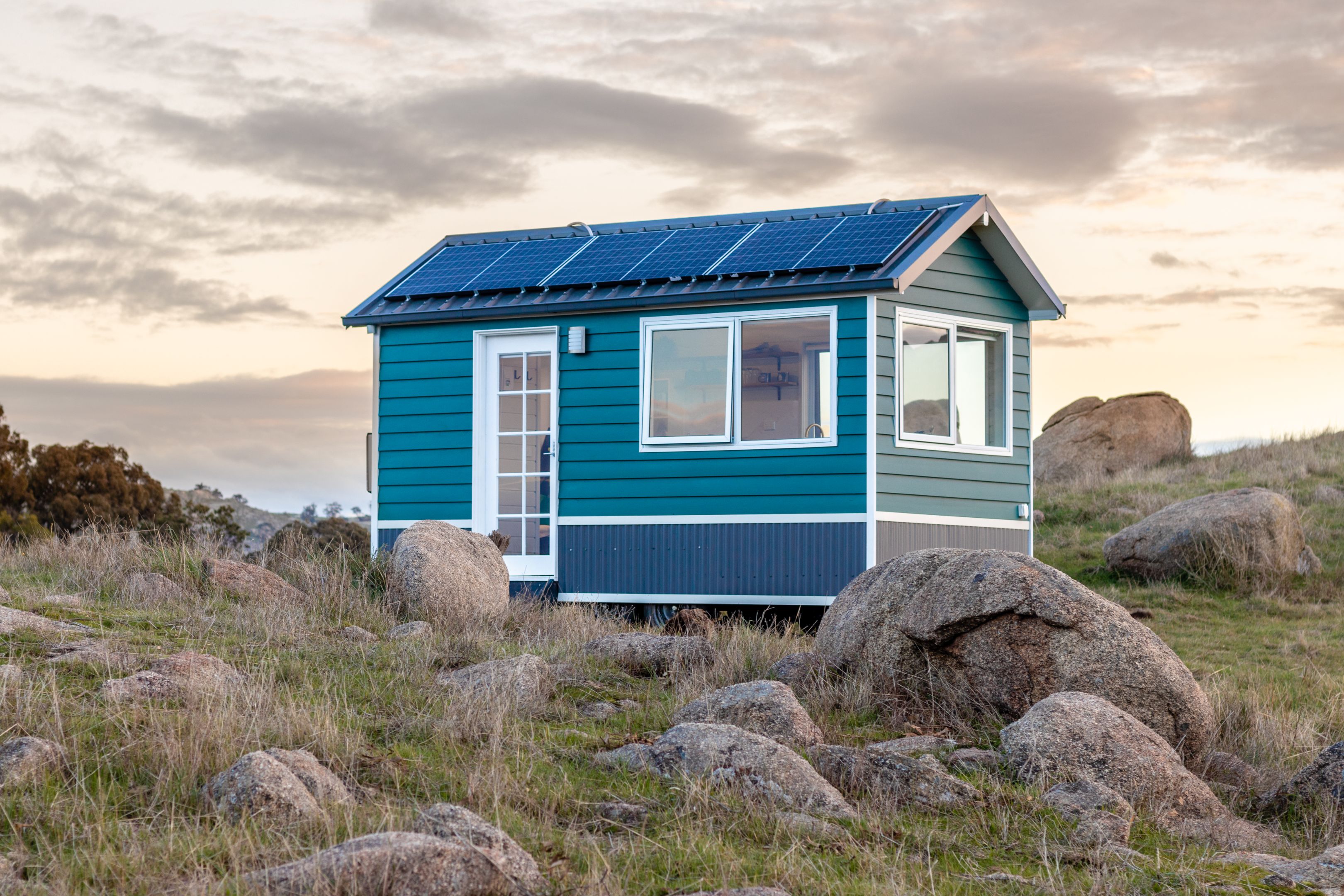 Aimee Stanton's Tiny Stays Heathcote house A tiny blue cottage is nested among large rocks, green grass and a rich sunset