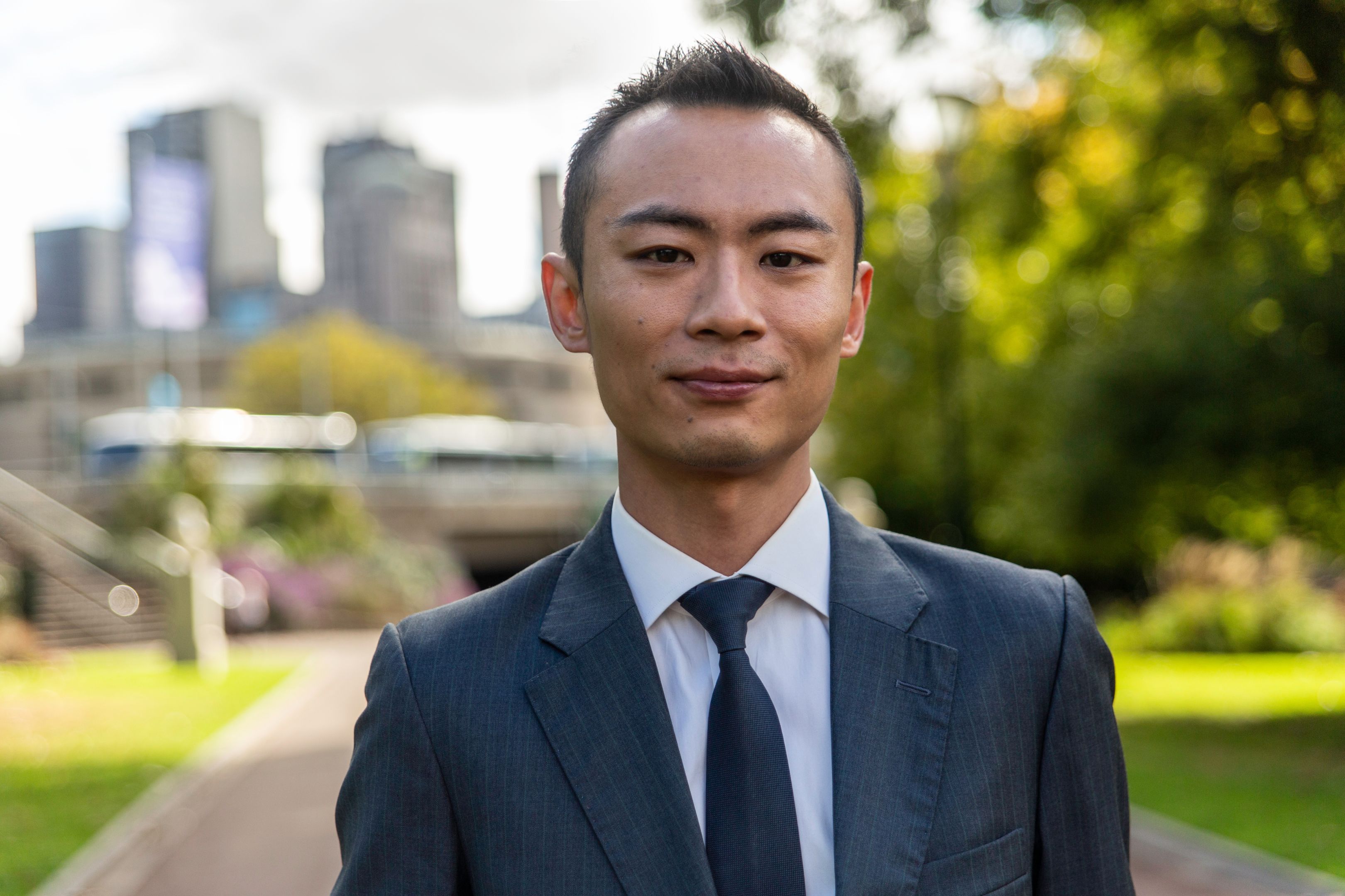 Christian Tang Close up of Christian Tang in suit attire with park greenery and Melbourne CBD blurred in background