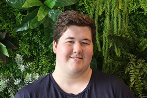 Rohan Gerrard A man wearing a navy t-shirt with brown hair smiles to camera surrounded by green foliage