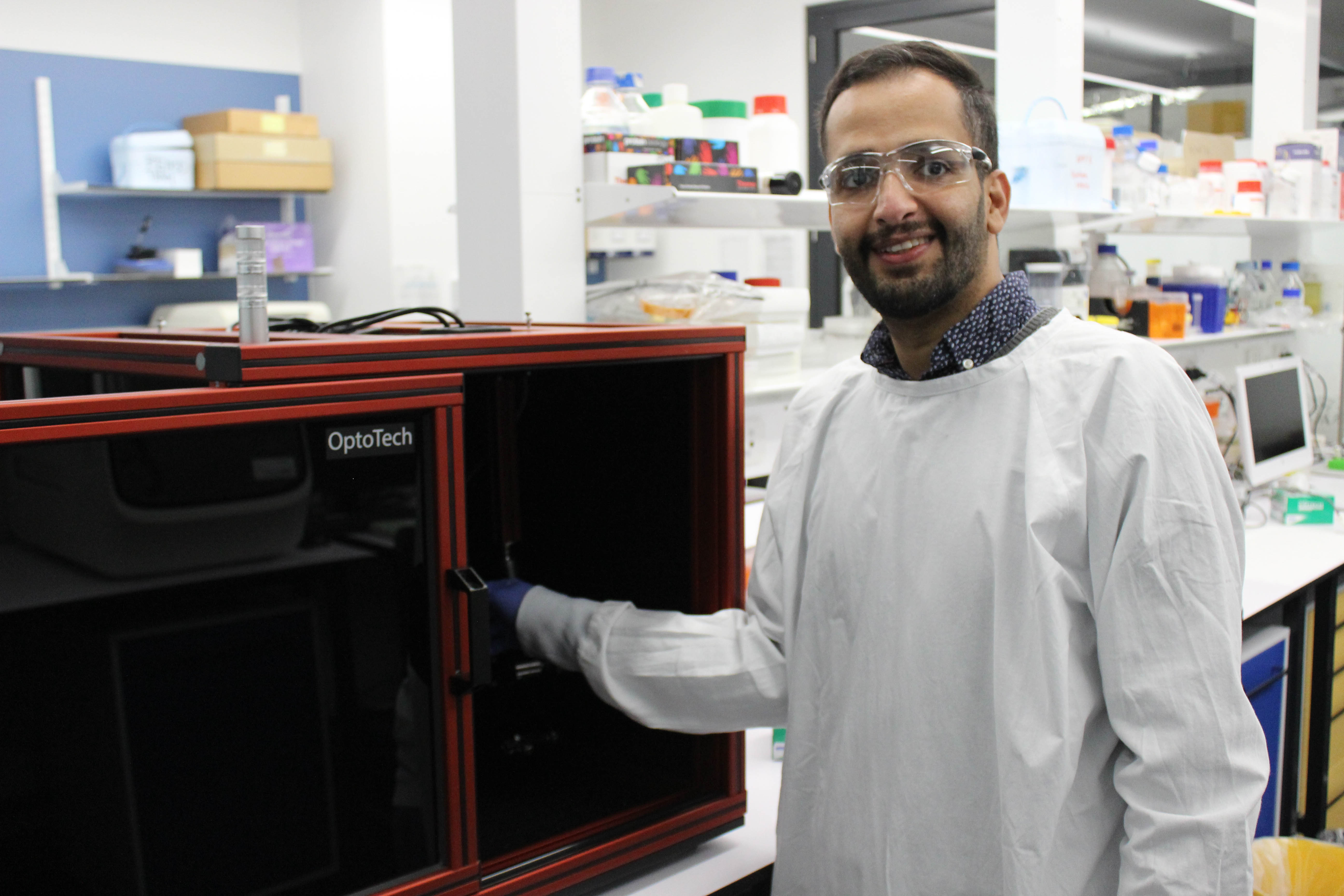 A man wearing a lab coat and blue plastic gloves stands in a laboratory with white shelves lined with an assortment of jars and boxes. On a table beside him is a glass and lightweight metal laser enclosure in the shape of a large cube like a microwave