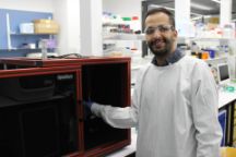 A man wearing a lab coat and blue plastic gloves stands in a laboratory with white shelves lined with an assortment of jars and boxes. On a table beside him is a glass and lightweight metal laser enclosure in the shape of a large cube like a microwave