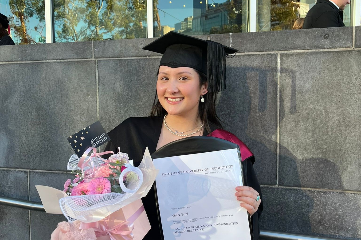 Student on her graduation day in her graduation hat and gown holding some flowers and her certificate