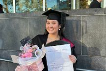 Student on her graduation day in her graduation hat and gown holding some flowers and her certificate