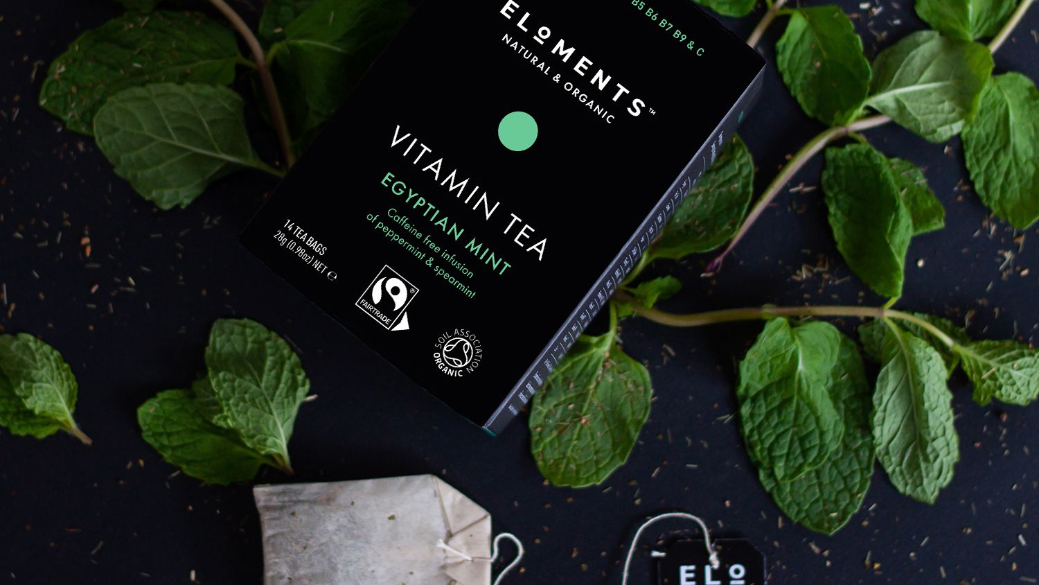 Eloments Vitamin Tea A black box of Eloments Vitamin Tea 'Egyptian Mint' flavour sits on a black stone background atop fresh mint. One tea bag is displayed on the right hand side.