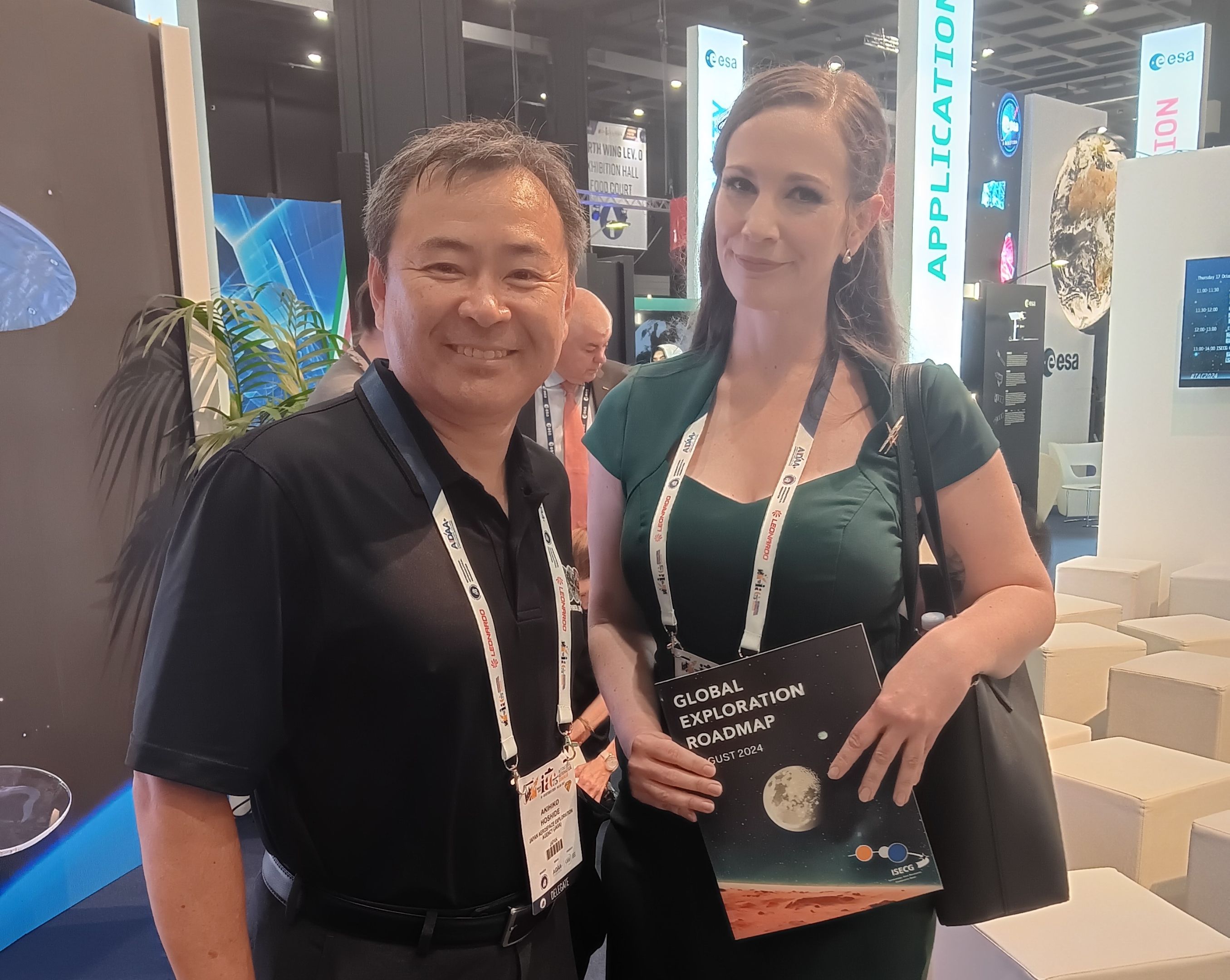 Erin Rose with Akihiko Hoshide at the IAC 2024 A women in a green dress poses beside a man in a black shirt. They are both smiling with lanyards around their necks
