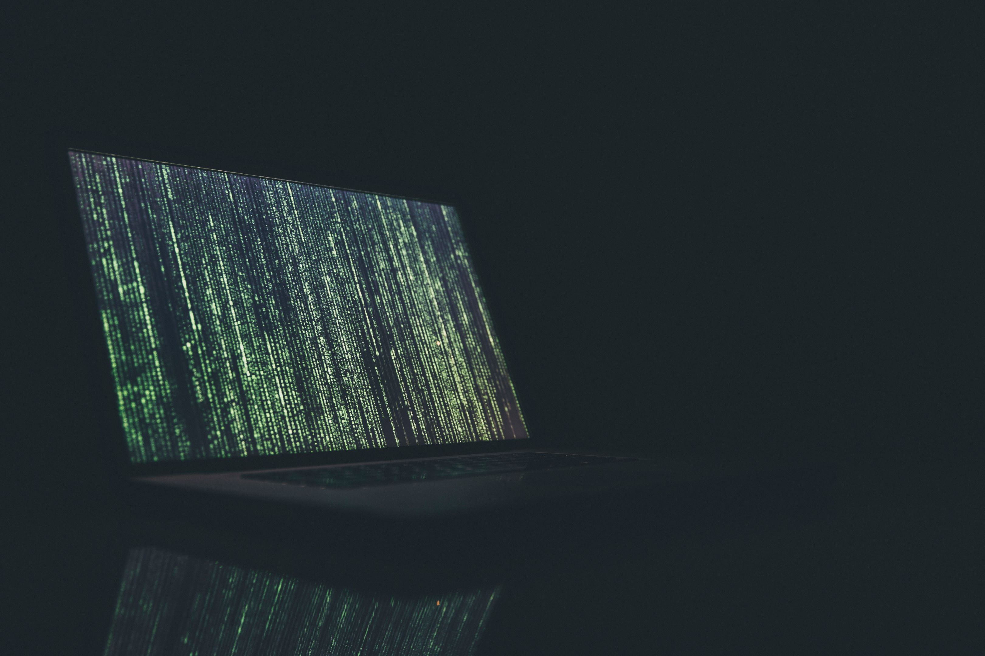 A laptop sits on a black glossy bench in a dark room. On it's screen is green and black code. The screen illuminates the room with a greenish light. 