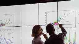 Business men in a dark room standing in front of a large data display analysing graphical data.